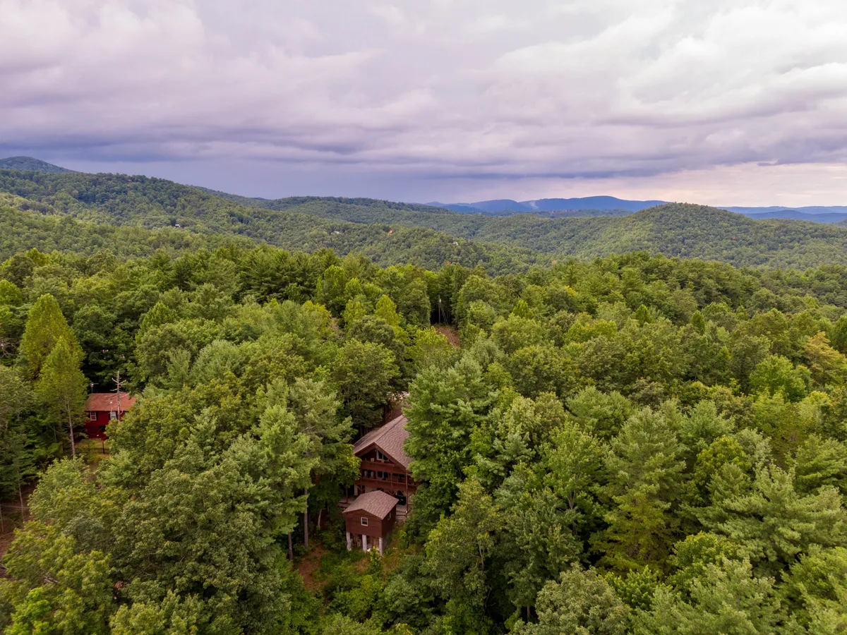 This image features a classic wooden cabin nestled among tall trees with a dramatic mountain range forming the backdrop. The scene is tranquil, capturing the lush natural surroundings and the promise of mountain adventures just outside the door.