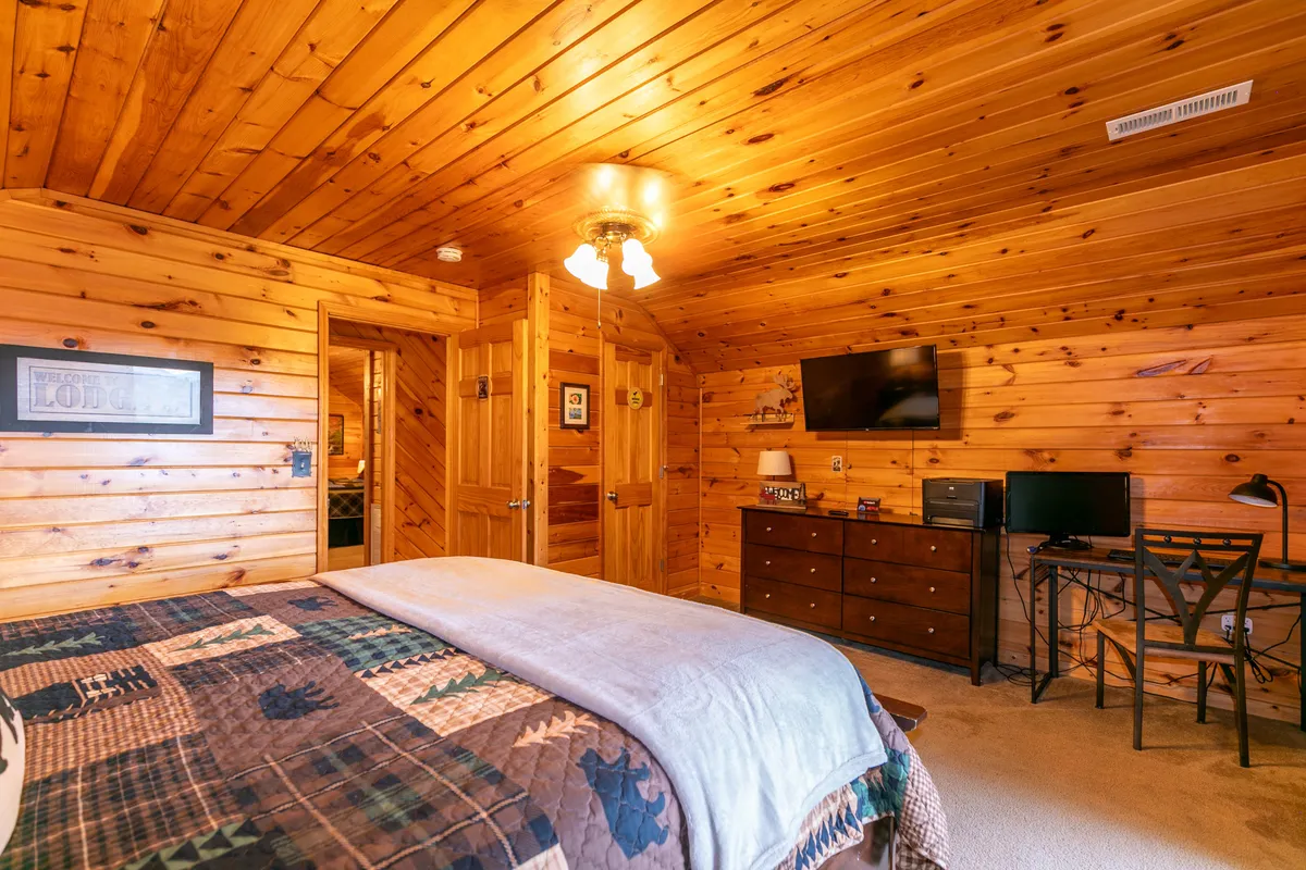 A cozy bedroom with a quilt-topped bed, wooden walls, a writing desk (complete with a chair), and a television. Sunlight filters through, inviting guests to relax, work, or unwind in privacy.