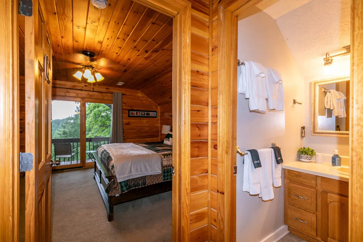 A serene bedroom featuring a stylish bed with a decorative throw, set against rustic wooden beams and large windows that frame the lush outdoor view. The space is flooded with natural light, creating a tranquil atmosphere ideal for relaxing.