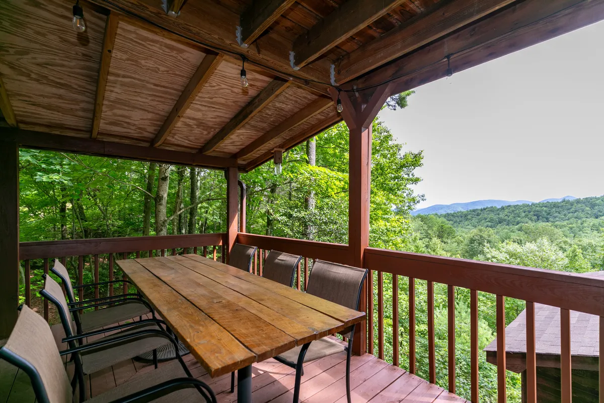 An inviting wooden deck set up for outdoor dining, surrounded by lush greenery and offering breathtaking mountain views. The large table suggests group gatherings while the scenery emphasizes the outdoor connection.
