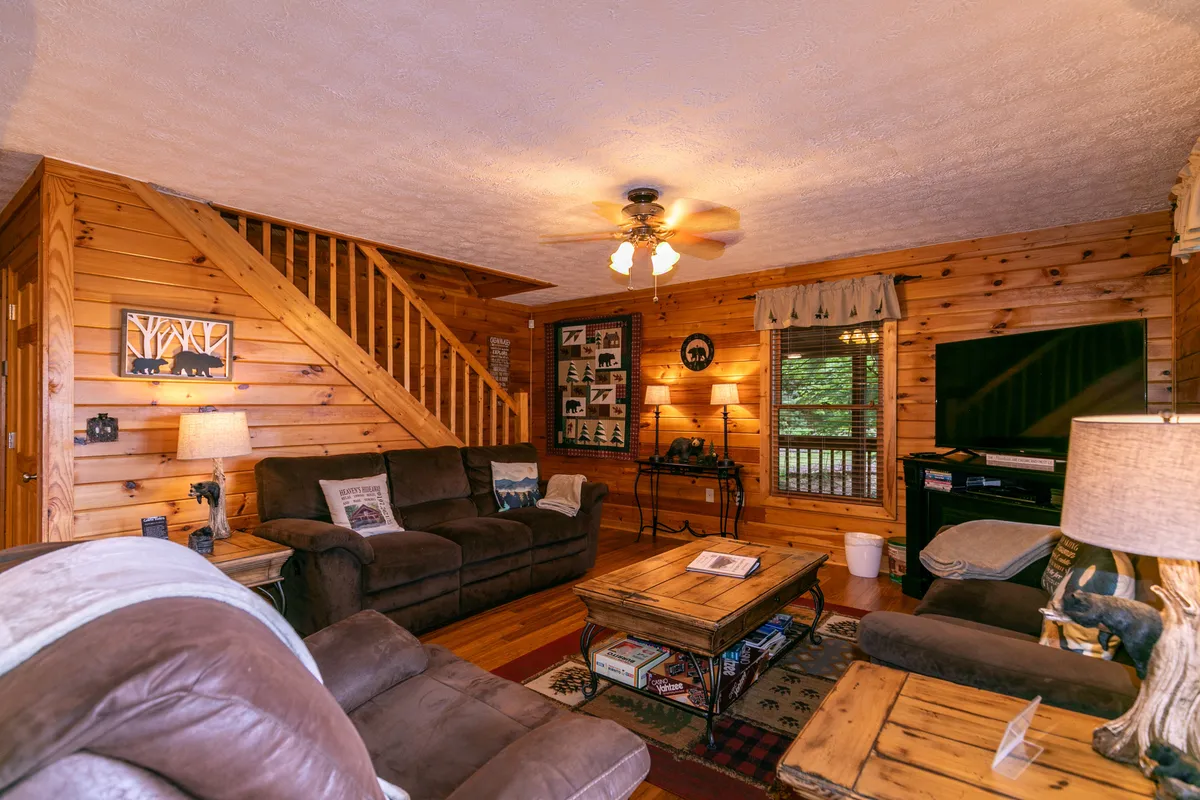 A warm and inviting living room with comfortable sofas arranged around a coffee table, a TV mounted on the wall, a decorative wooden staircase, and wall art adding to the cozy ambiance. The space is well-lit and designed for gatherings, relaxation, and entertainment.