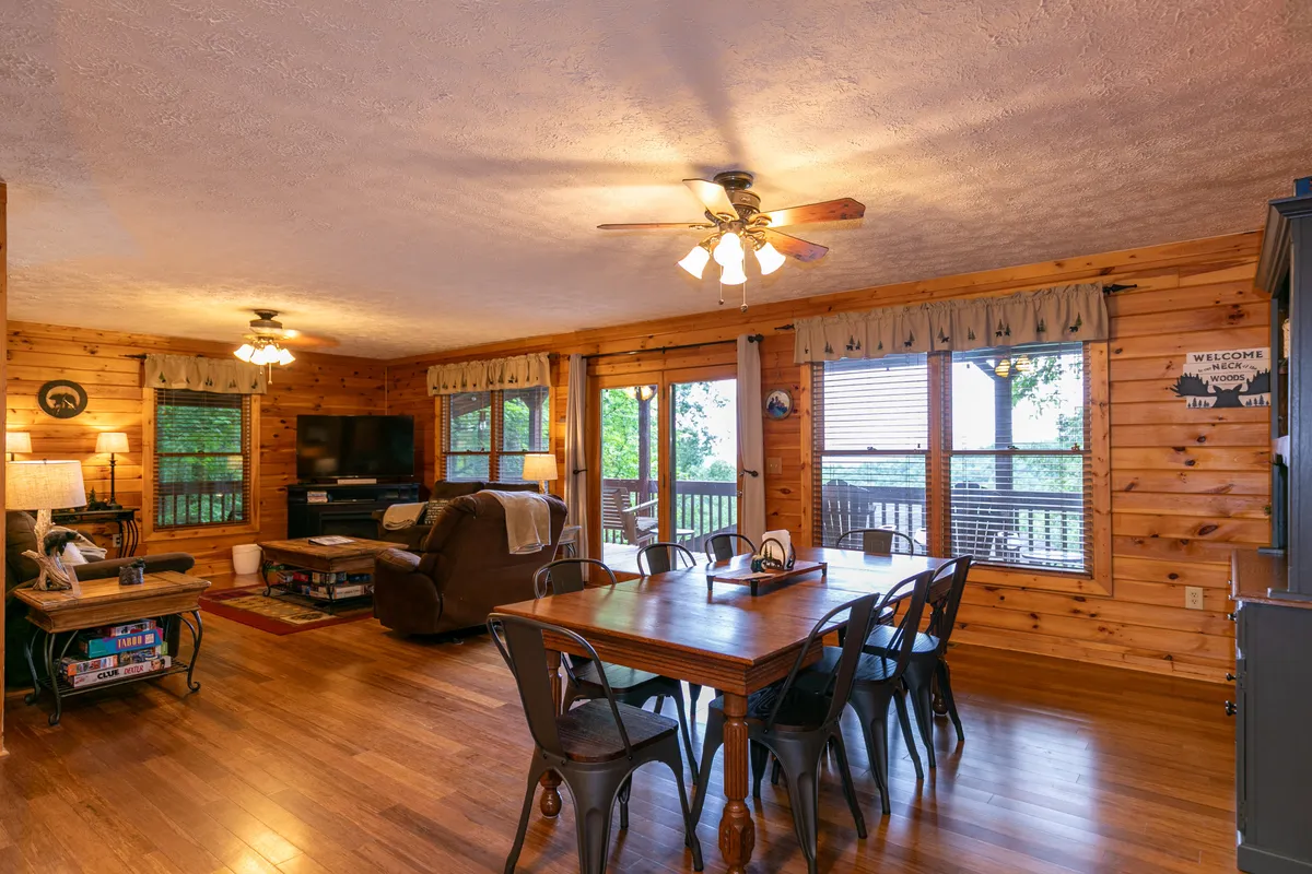 An open and inviting scene showing a dining table adjacent to a sofa and a television, with large windows that allow natural light to stream in. The setup promotes seamless transitions from dining to relaxing, with views to the outside and a cozy atmosphere for movie nights or family time.