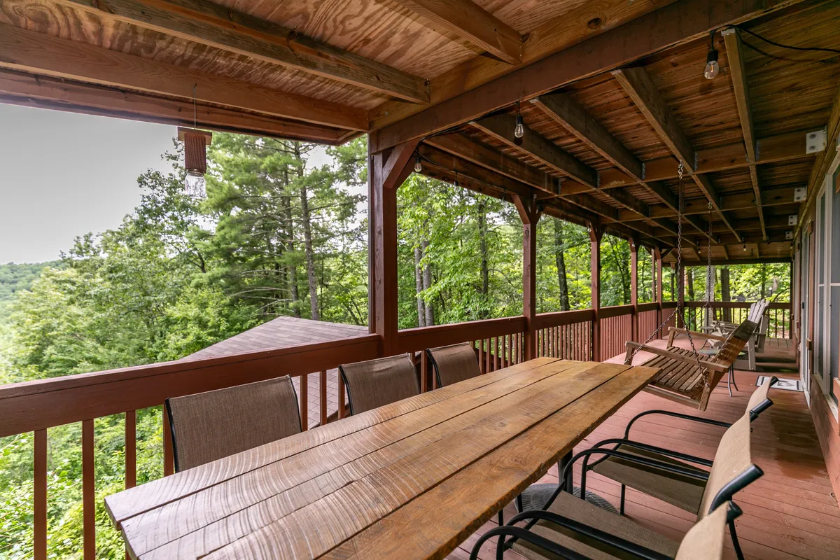 A spacious wooden deck set for communal dining, bordered by thick greenery and illuminated by hanging light fixtures. The scene suggests relaxation, group meals, and seamless indoor-outdoor living.