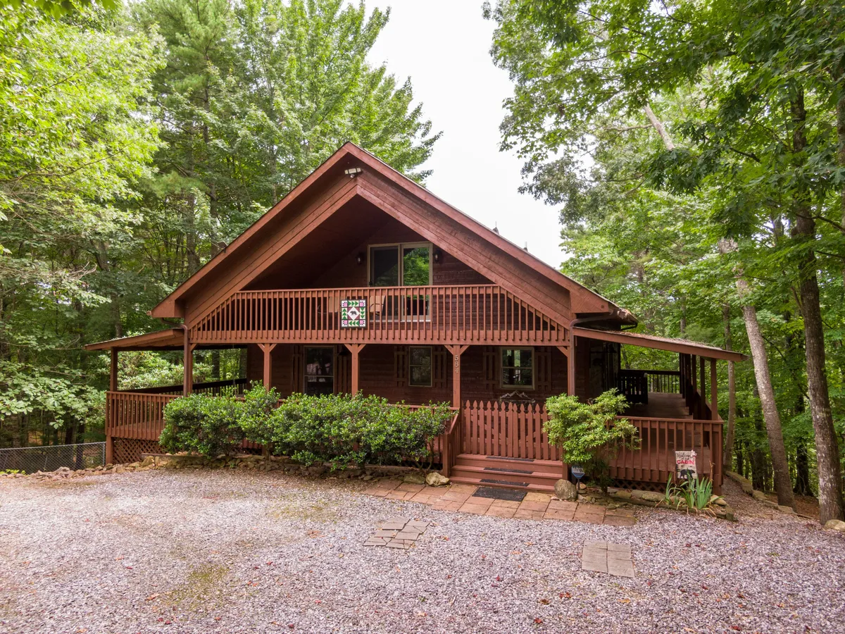 The hero image showcases the welcoming exterior of a charming log cabin, nestled amidst lush greenery. A spacious wooden porch wraps around the front, ideal for relaxing and taking in the surrounding natural beauty. Stone pathways lead up to the front steps, inviting guests to enter and explore. The overall scene radiates tranquility and warmth, suggesting opportunities for family gatherings, morning coffee on the porch, and leisurely outdoor activities.