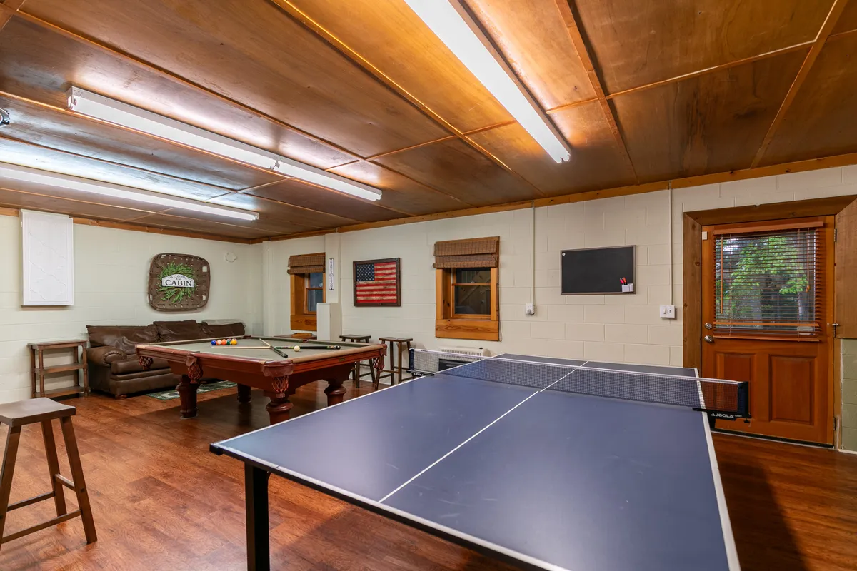 This inviting interior scene features both a ping pong table and a pool table, set in a cozy, wood-accented recreational room. A comfortable sofa sits nearby, and an American flag adds character. The setup is ideal for game nights, casual hangouts, or friendly competitions after hours.